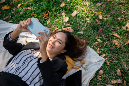 A carefree and happy Asian businesswoman is using her smartphone while lying on a picnic mat in a...
