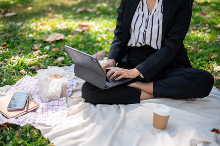 A positive and attractive Asian businesswoman remote working at a park, working on her digital tablet on a picnic mat. cropped shotの写真素材