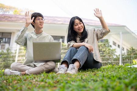Two cheerful and friendly Asian university students are sitting on the grass in a campus park,...