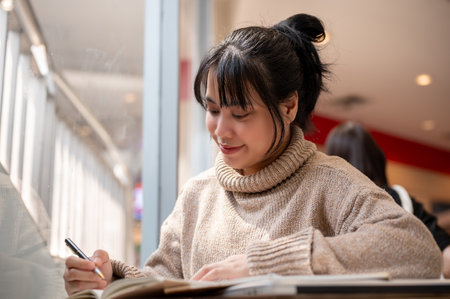 A beautiful and positive Asian woman in a comfy knitted sweater is keeping her diary or making a list of her ideas while working remotely at a cafe.の写真素材