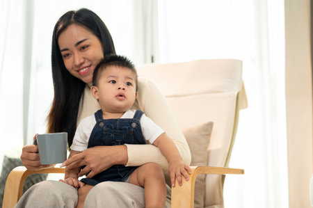 A young Asian mom is having her morning coffee and sitting on a lounge chair with her little son in the living room.の写真素材