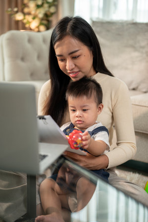 A positive Asian businesswoman mom and her baby boy are reading documents together in the living room, taking care of her baby while working from home.の写真素材