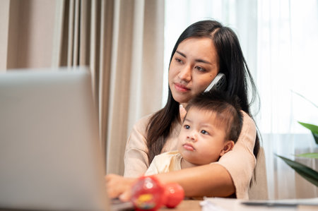A busy Asian businesswoman mom is multitasking, talking on the phone with her client, focusing on her laptop, and taking care of her little son at home. work from home and mom's life conceptsの写真素材