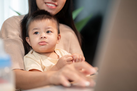 A positive Asian mom is taking care of her baby boy while working on her laptop computer, working remotely from home.の写真素材
