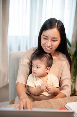 A positive Asian mom is taking care of her baby boy while working on her laptop computer, working remotely from home.の写真素材