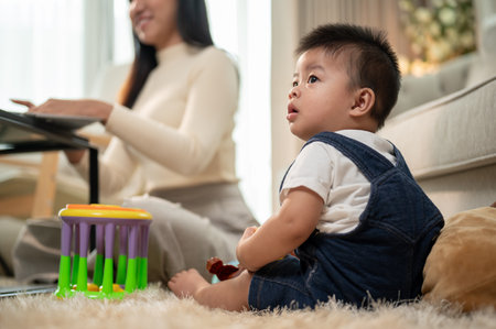 A cute Asian baby boy is sitting on a carpet, playing with toys in the living room while his mom is...