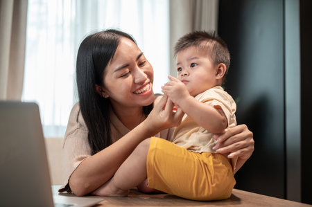 A happy Asian mom is playing and feeding her little son with snacks while working in her home office. work from home and mom's life conceptsの写真素材