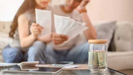 A close-up image of a jar of coins on a coffee table with a blurred background of a stressed couple...
