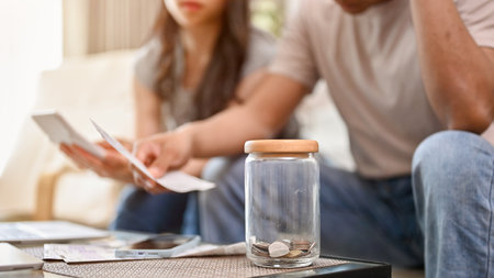 A close-up image of a jar of coins on a coffee table with a blurred background of a stressed couple...