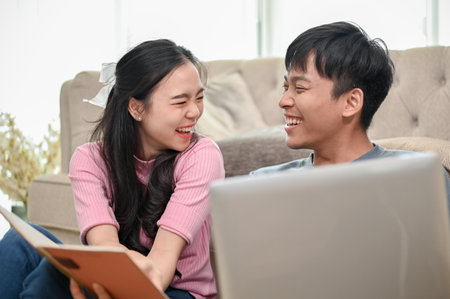 A cheerful young Asian couple is laughing and enjoying talking while working at home together, using a laptop and reading a book. spouses, fiancÃ©, boyfriend and girlfriend, husband and wifeの写真素材