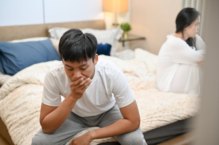 A sad, unhappy Asian husband in pajamas is sitting separately with his wife on bed after their argument. relationship problems, misunderstanding, marriage conflictの写真素材