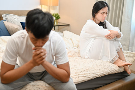 A sad, unhappy young Asian wife in pajamas is sitting separately with her husband on bed after their argument. relationship problems, misunderstanding, marriage conflictの写真素材