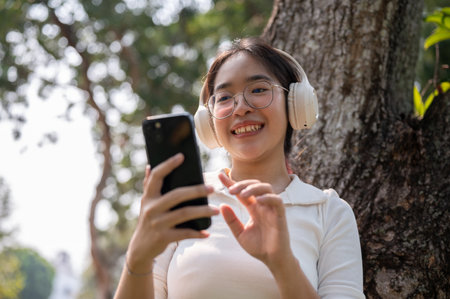 A beautiful young Asian woman is wearing her headphones and using her smartphone under the tree in a...