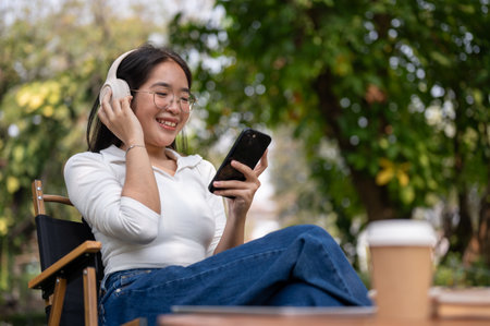 A relaxed and happy Asian woman is sitting in a green backyard, listening to music on her headphones, and using her smartphone. people and lifestyle conceptsの写真素材