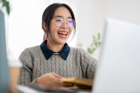 A cheerful young Asian female college student is studying online in a cafe co-working space, participating in an online class. people and education conceptsの写真素材