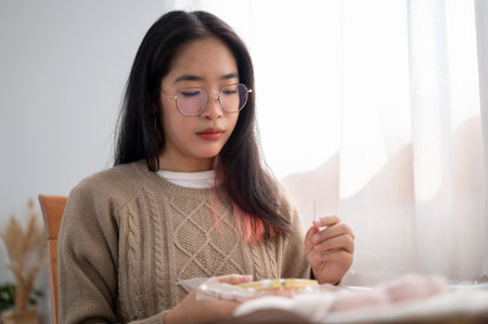 A young Asian woman wearing eyeglasses is focusing on threading a pattern on an embroidery frame, hand sewing on cloth, while sitting at a table by the window indoors. Handmade needleworkの写真素材