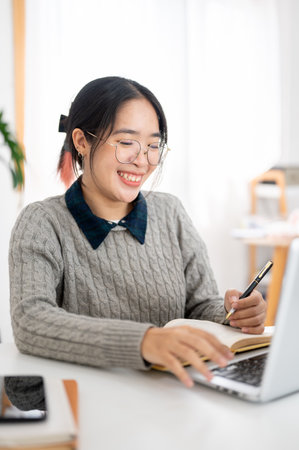 A cheerful young Asian female college student is studying online in a cafe co-working space,...