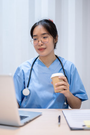 A professional and experienced Asian female doctor is sipping coffee while reading medical cases on laptop in her office.の写真素材