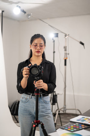 A professional, experienced Asian female photographer is adjusting her DSLR camera on a tripod stand, preparing shooting equipment in the studio.の写真素材