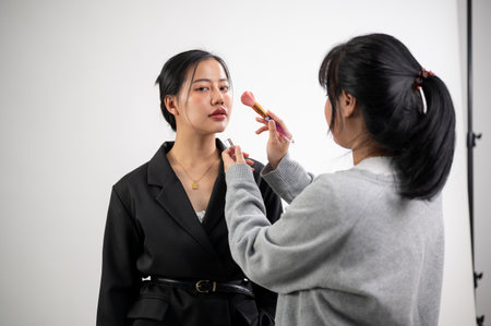 A beautiful young Asian female model is having makeup applied by a female makeup artist on a set, preparing for a fashion magazine photoshoot in the studio with professional lighting equipment.の写真素材