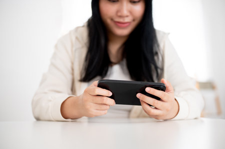 A cropped image of a young Asian woman in casual wear is using her smartphone, watching a video or...