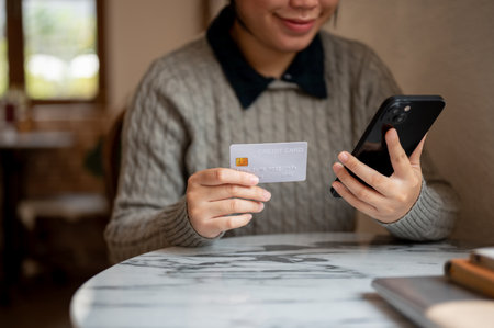 A cropped shot of an Asian woman holding her smartphone and a credit card, sitting at a table indoors, shopping online, registering a credit card account on a shopping website, or transferring money.の写真素材