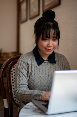 A beautiful, positive young Asian woman in casual wear is focusing on her work on her laptop...