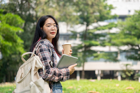 A positive young Asian female college student in glasses and casual wear carrying her backpack, digital tablet, and a coffee cup standing in a campus park on a sunny day.の写真素材