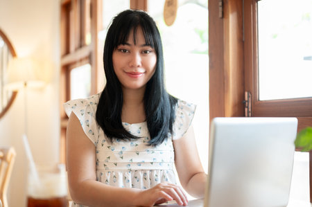 A beautiful young Asian woman sitting in a coffee shop with her laptop computer, working remotely, smiling at the camera. people and technology conceptsの写真素材