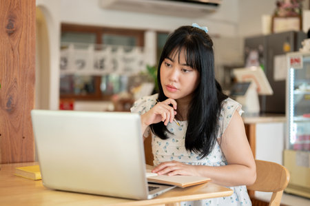A serious, thoughtful young Asian woman in casual clothes is focusing on her work, reading an online article on the website, looking at her laptop screen, working remotely at a coffee shop.の写真素材