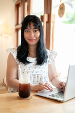 A beautiful young Asian woman sitting in a coffee shop with her laptop computer, working remotely, smiling at the camera. people and technology conceptsの写真素材