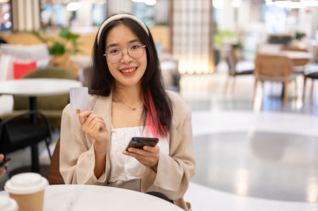 A happy young Asian woman is showing her credit card to the camera while sitting in a restaurant in...