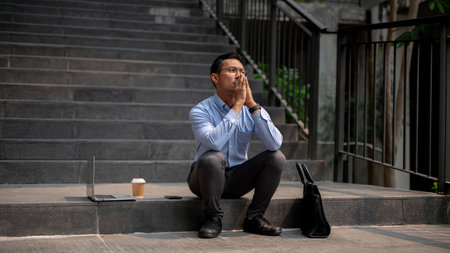 An Asian millennial businessman sits on outdoor steps with a laptop, a briefcase, and a coffee cup...