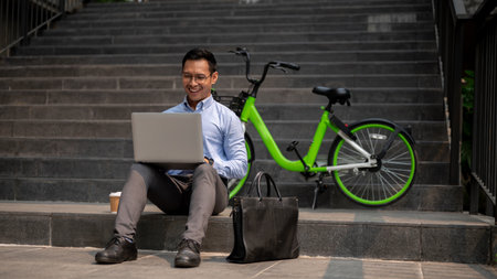 A confident Asian businessman sits on steps with a green bicycle beside him, working on a laptop with a smile, working remotely from the city. businesspeople and modern work culture conceptsの写真素材