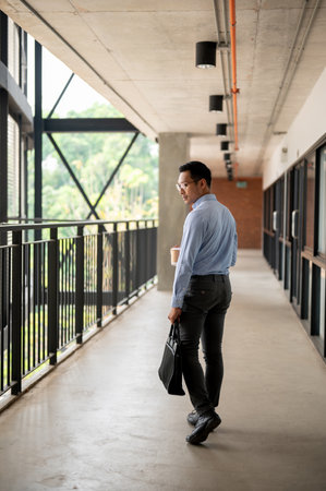 A determined, confident Asian millennial businessman walking along an indoor corridor in the company building, holding a coffee cup and a briefcase. businesspeople conceptの写真素材