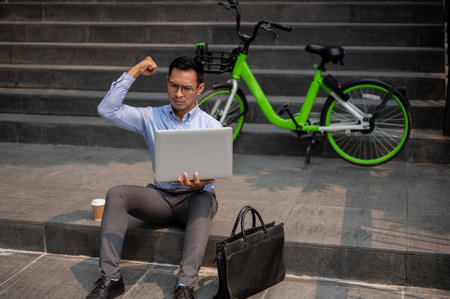 A determined Asian millennial businessman sits on steps outside with his laptop, showing his fist, celebrating a successful achievement or feeling empowered by progress.の写真素材