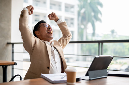 A cheerful Asian businessman in a beige suit and glasses raises his arms triumphantly while sitting at a table in a building corridor, celebrating success or good news received on his digital tablet.の写真素材