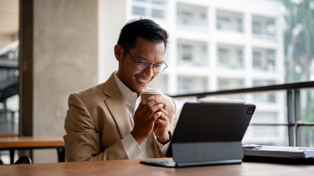 A happy Asian businessman in a beige suit and glasses smiles as he holds a disposable coffee cup while looking at a digital tablet, sits at a table in a building corridor, feeling relaxed during work.の写真素材