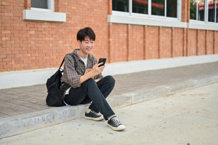 A positive, happy young Asian male college student with a backpack sits on the street near the campus building using his smartphone. university life, lifestyle, wireless technologyの写真素材
