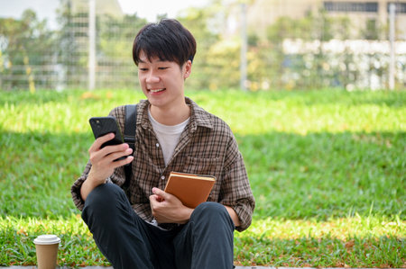 A cheerful young Asian male college student sits on the street in the campus park using his phone, reading text, checking messages, or chatting with his friends.の写真素材