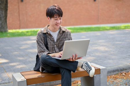 A young Asian male college student using his laptop computer on a bench in the campus park. university life and wireless technology conceptsの写真素材