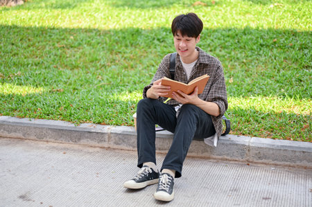 A positive, handsome young Asian male college student sits on the street in the campus park reading a book, enjoying his outdoor leisure. university life, lifestyle, hobbyの写真素材