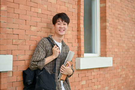 A smiling young Asian male college student in casual wear stands by a brick wall on campus, carrying a backpack, holding a laptop and books, and looking at the camera.の写真素材