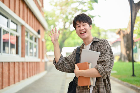 A friendly, positive young Asian male college student in casual wear, stands outdoors on campus, carrying a backpack, holding a laptop and books, waving his hand and smiling at the camera.の写真素材