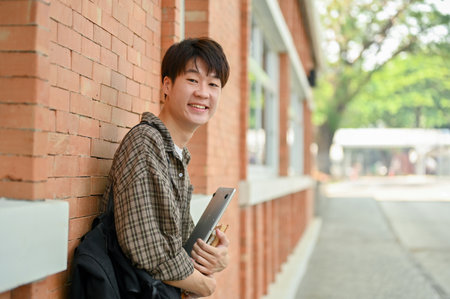 A smiling young Asian male college student in casual wear stands by a brick wall on campus, carrying a backpack, holding a laptop and books, and looking at the camera.の写真素材