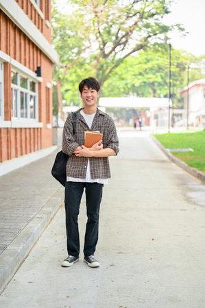 A smiling, positive young Asian man college student in casual wear, carrying a backpack, stands in front of a brick building on campus, holding books.の写真素材