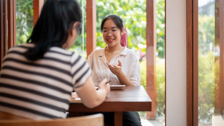 A positive Asian woman is having an informal meeting with her client at a coffee shop, talking and explaining some details. A college student meeting her friend at a cafe, doing homework together.の写真素材