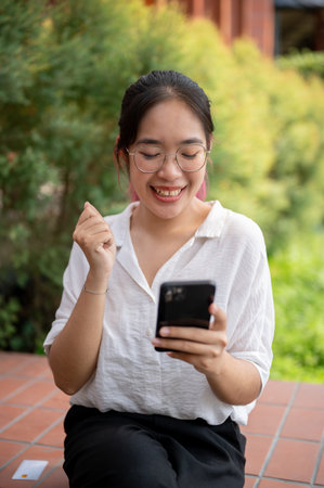 A young cheerful Asian woman in casual wear and glasses is looking at her smartphone screen with a happy face, using her smartphone while sitting on a bench in a park on a sunny day.の写真素材