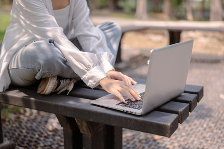A cropped image of an Asian woman in casual wear sits on a bench in a public park and uses her laptop computer, working remotely outdoors on a sunny day. lifestyle and wireless technology conceptsの写真素材