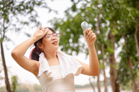 A young Asian woman wearing glasses and a tank top holds a small fan and wipes her sweat on her face...